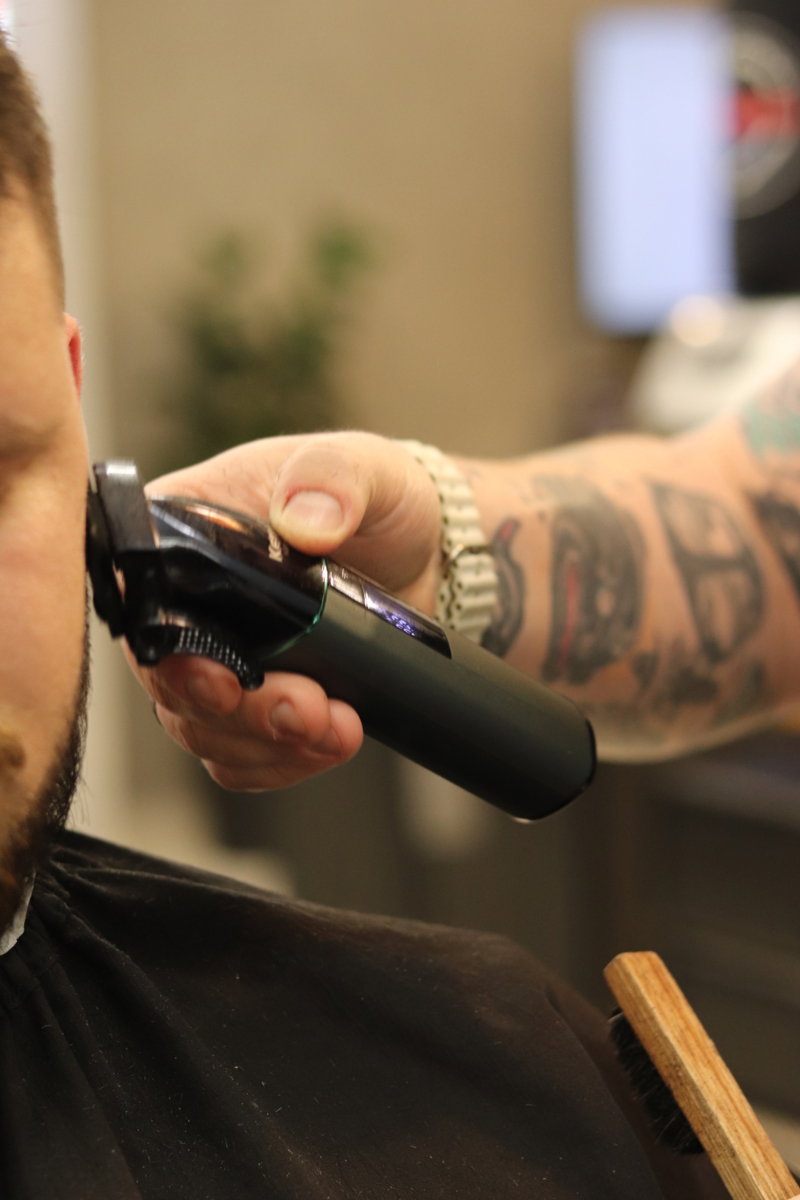 Máquina na nuca do cliente, com o antebraço tatuado do barbeiro
